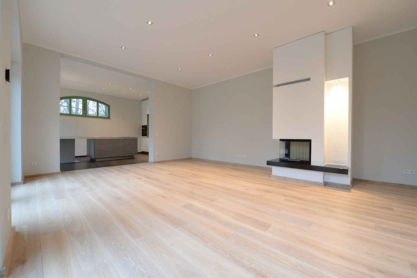 Empty living room with a pale board floor, an open fire in a white wall panel and a view through to the adjoining room.