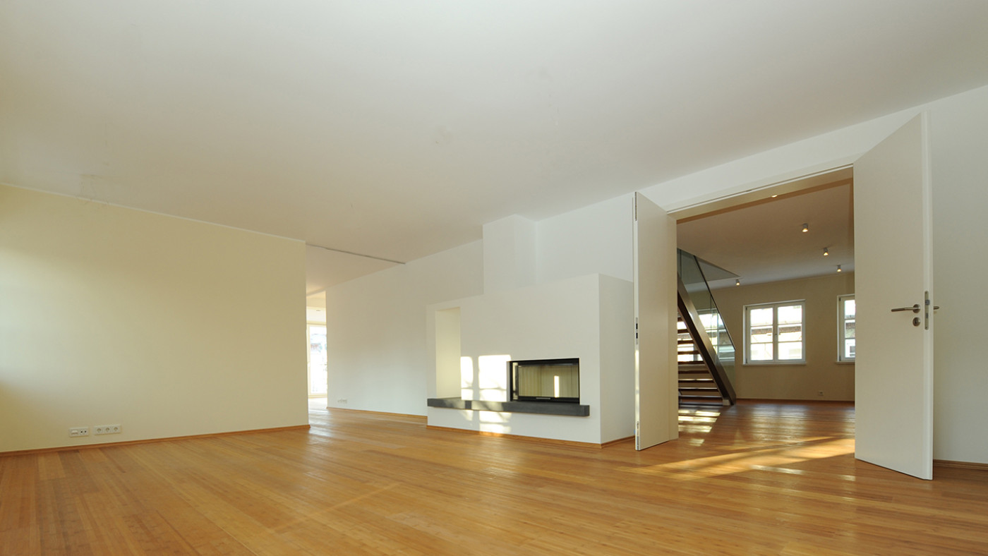 Empty refurbished living room with a parquet floor, a two-sided fireplace and a view towards the staircase.