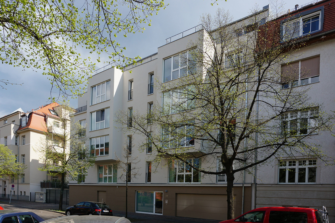The new-build section in pale render with balconies and a roof terrace between older buildings, a street with parked cars in front.