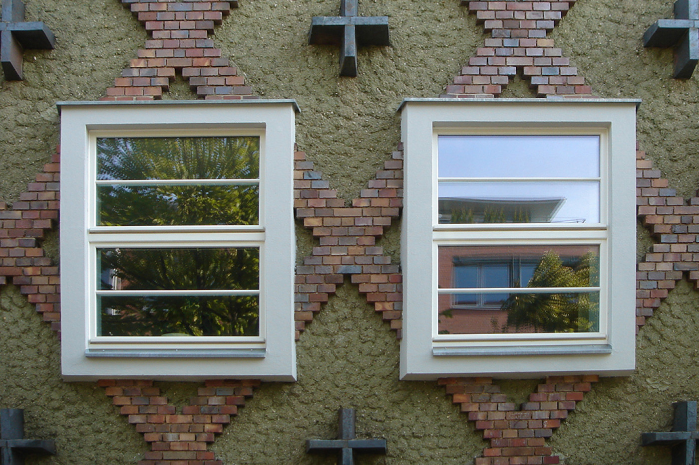 Two glazing-bar windows in the diamond-patterned fa&ccedil;ade with cross-shaped anchors, trees reflected in the panes.