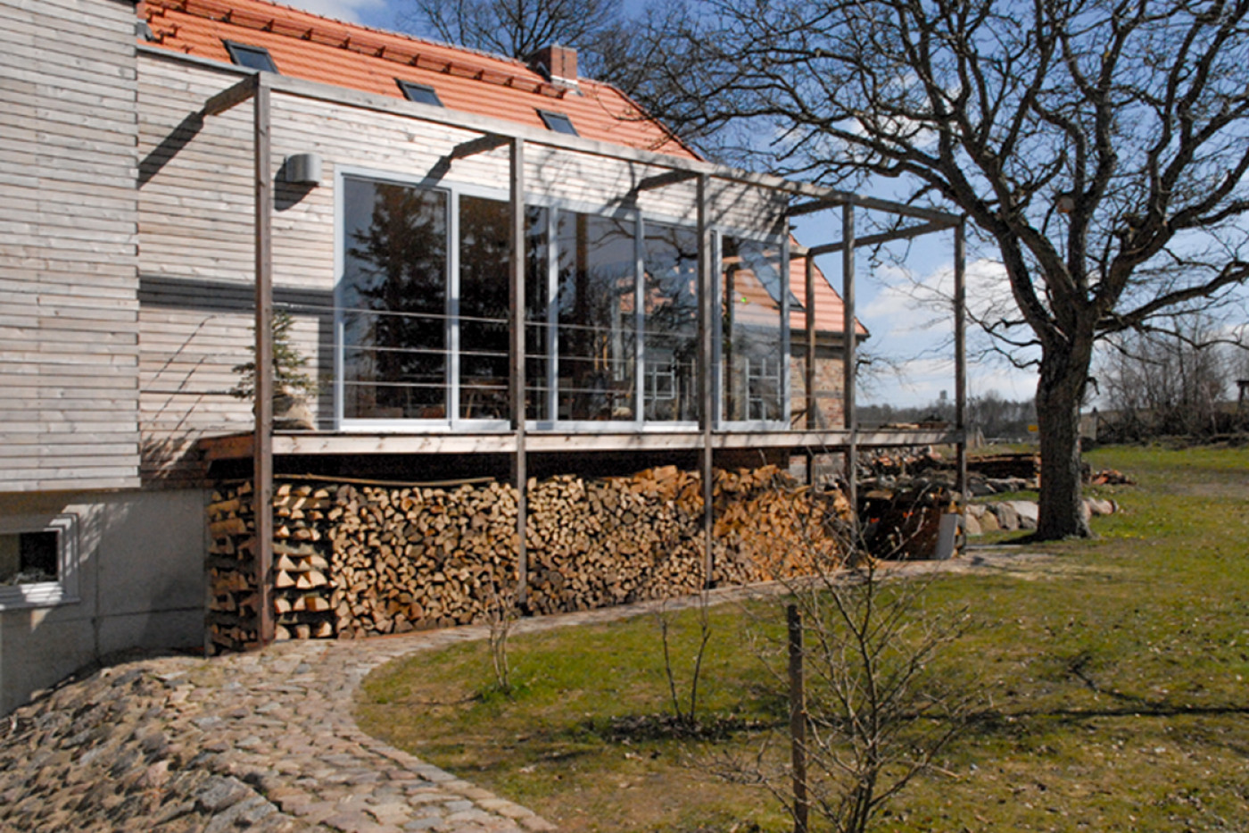Glazed extension with a timber pergola and terrace, stacked firewood beneath and a cobbled path in front.