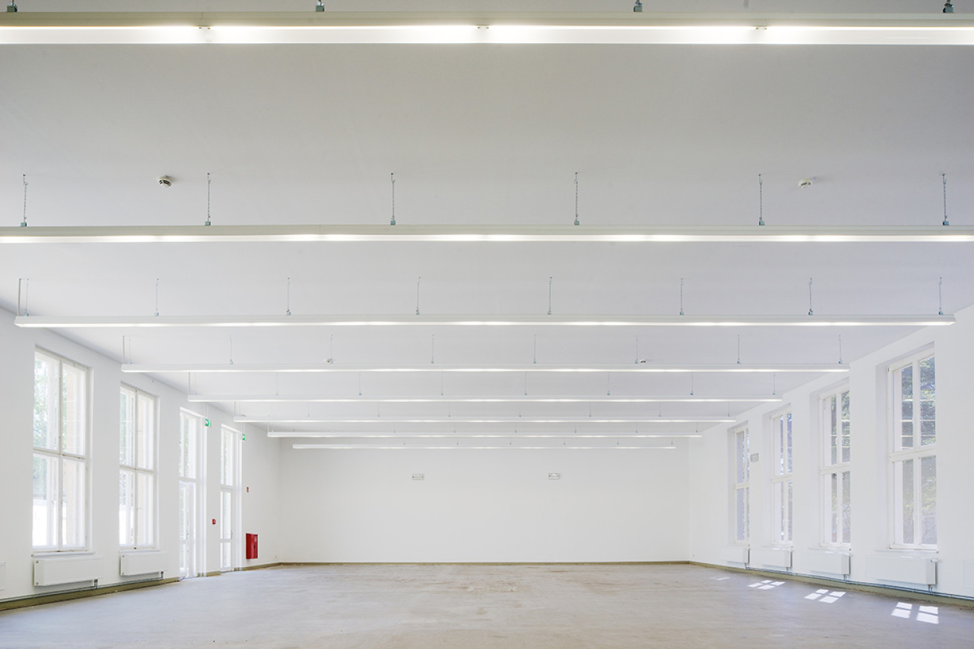 Empty white hall with downstand beams, lines of ceiling lights and tall glazing-bar windows along both sides.