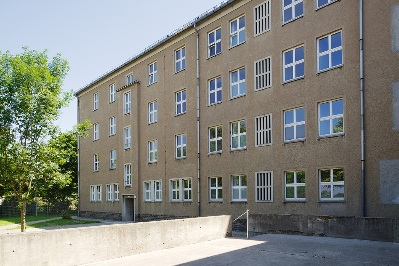 Four-storey rendered building with regular rows of windows and barred stair windows, a concrete retaining wall in front.