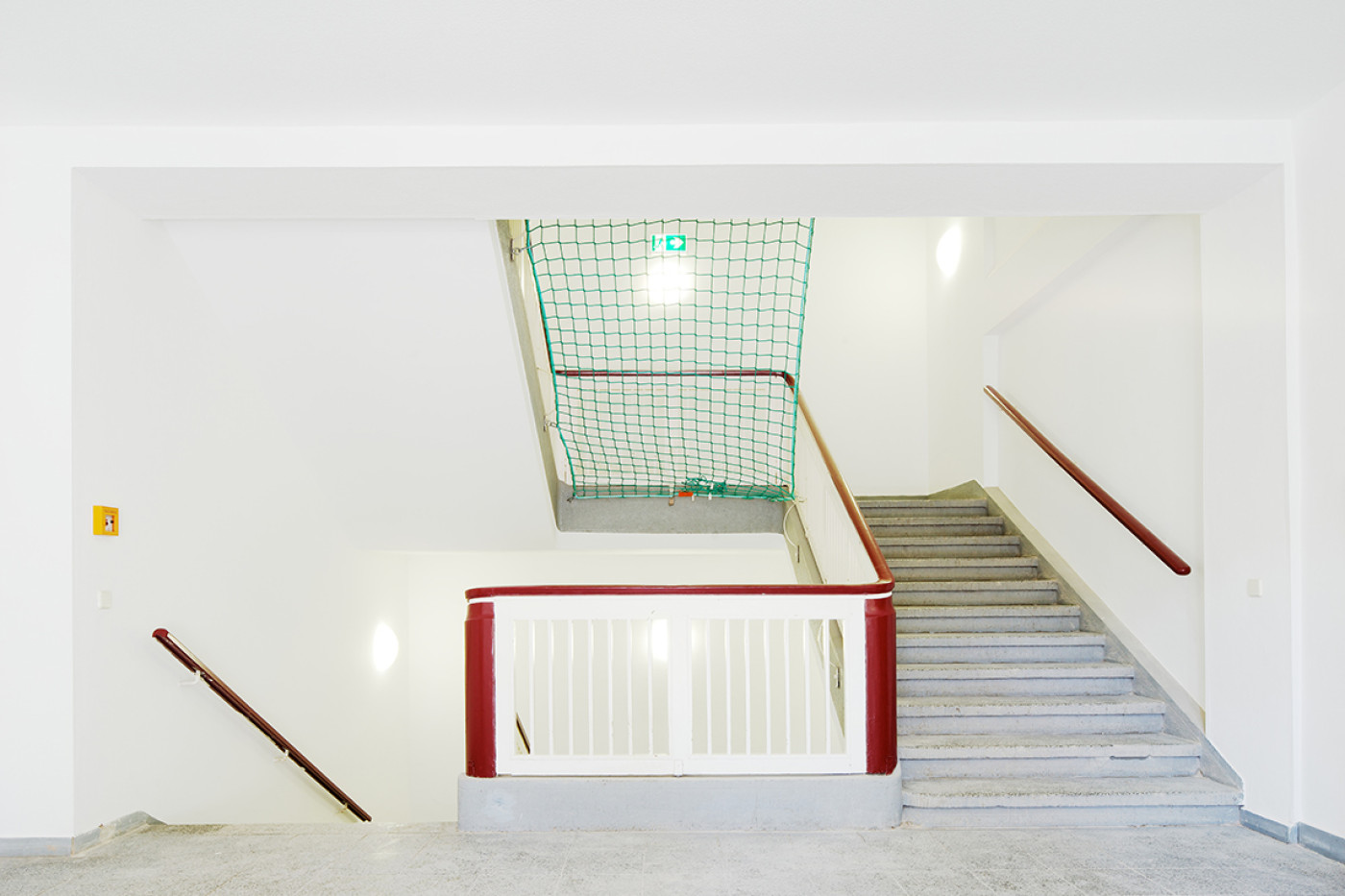 View into the stairwell with terrazzo steps, a red-painted balustrade and a green safety net in the void.