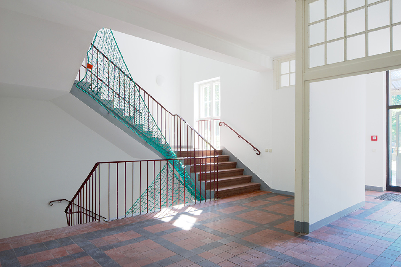 Stairwell with a red-brown tiled floor, dark red balustrade and a green safety net across the flight.