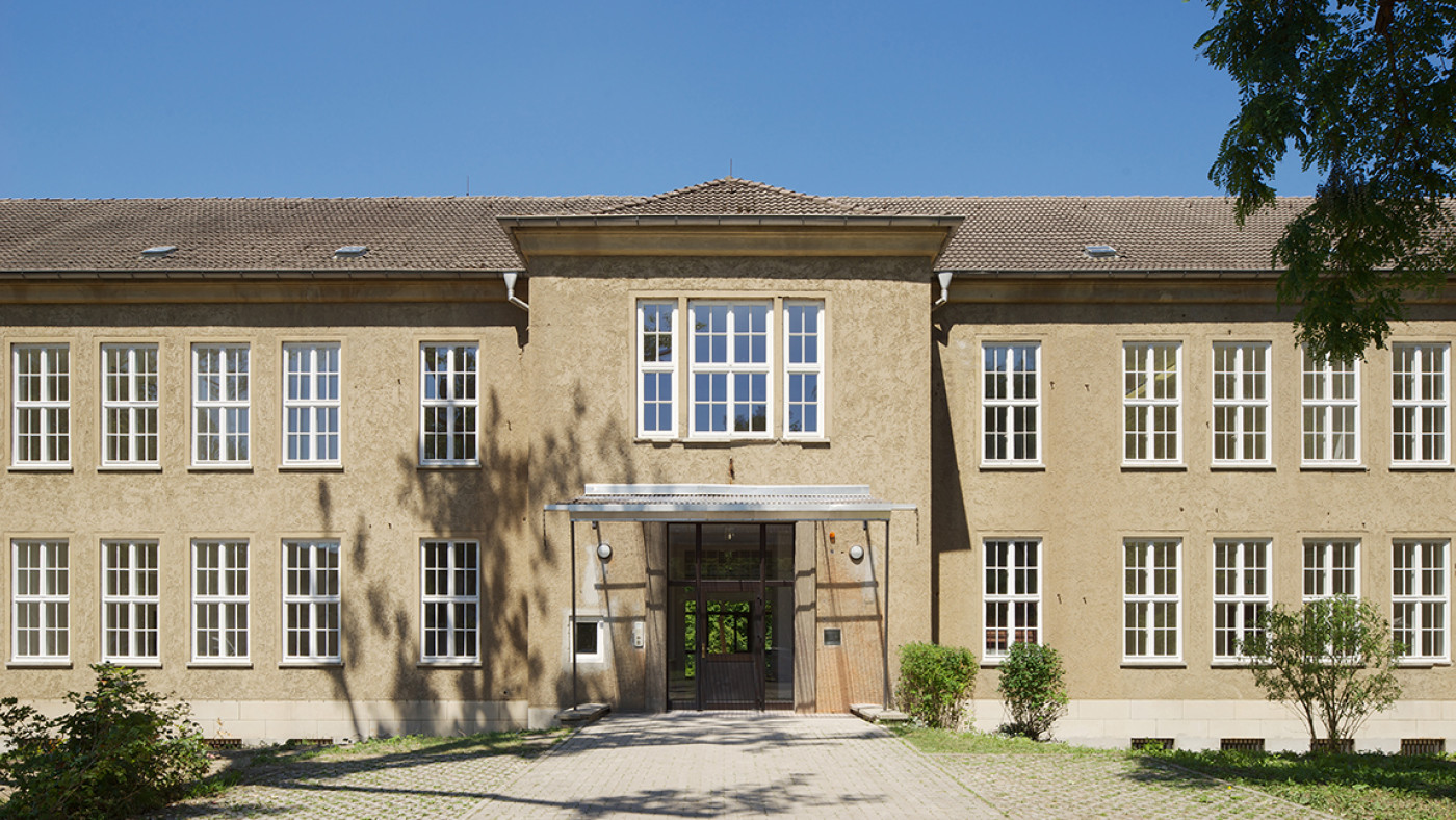 Two-storey rendered building with rows of windows and a covered entrance, the paved approach path in front.