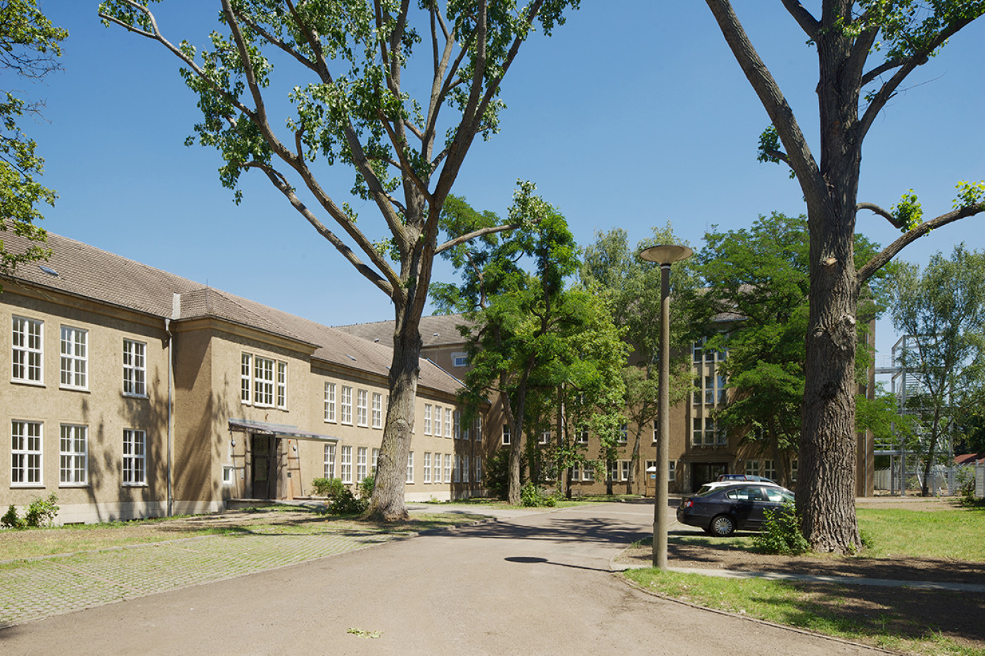 The long rendered building behind mature trees, an asphalt drive with a lamp post in front.