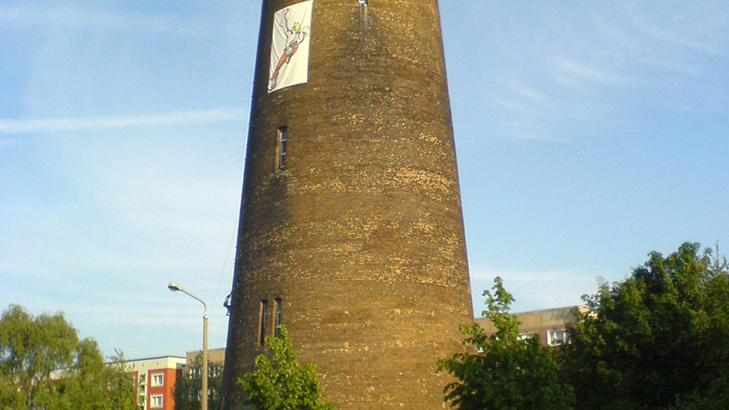 Runder Backsteinturm mit Klettersegel an der Fassade, umgeben von B&auml;umen.