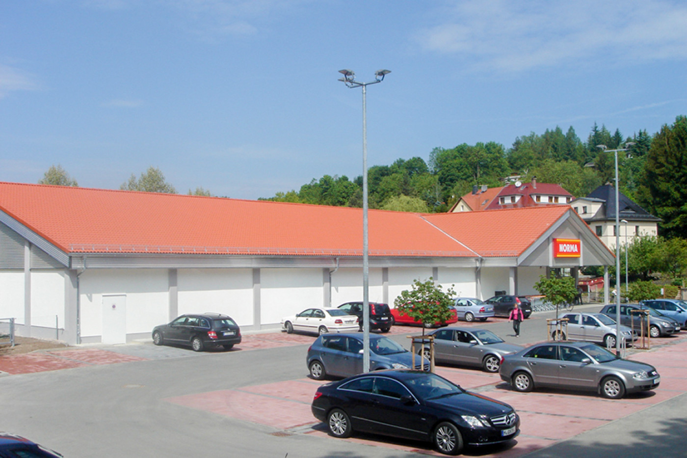 Eingeschossiger Nahversorger mit orangefarbenem Satteldach und Norma-Schriftzug, davor der Parkplatz mit Autos.