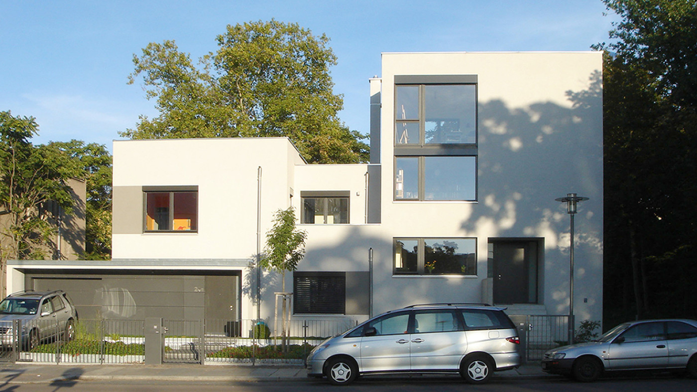 White semi-detached house with a flat roof and large dark-framed windows, an open carport on the left.