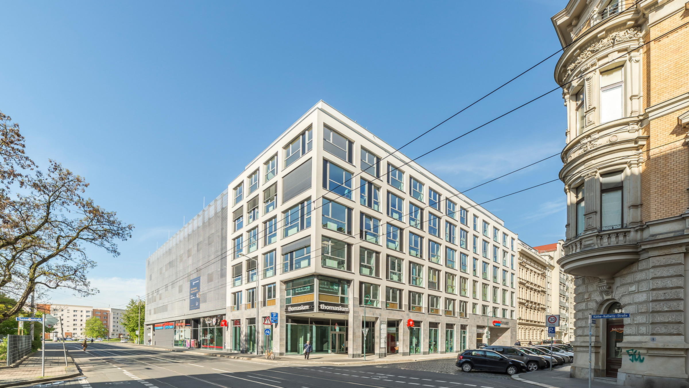 Corner building with a pale fa&ccedil;ade grid and retail floors, the multi-storey car park adjoining on the left.