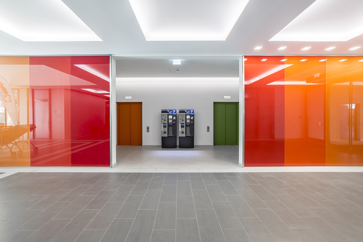 Passage with glass walls backed in red and orange, two doors in brown and green beyond and two machines between them.