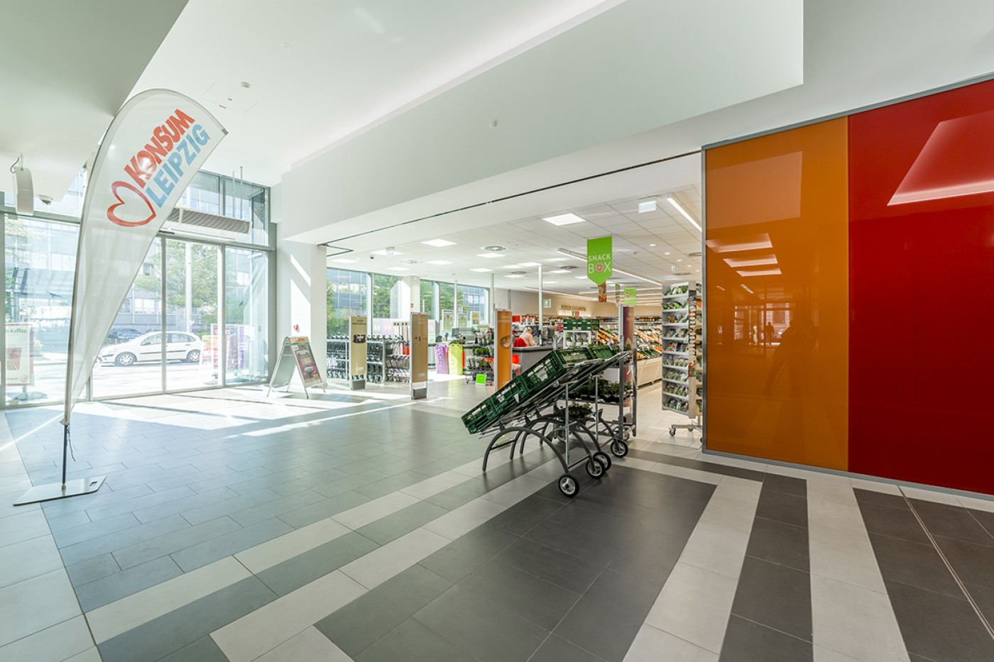 Entrance to the Konsum store with trolleys, an orange glass wall and a beach flag of the Leipzig Konsum co-operative.