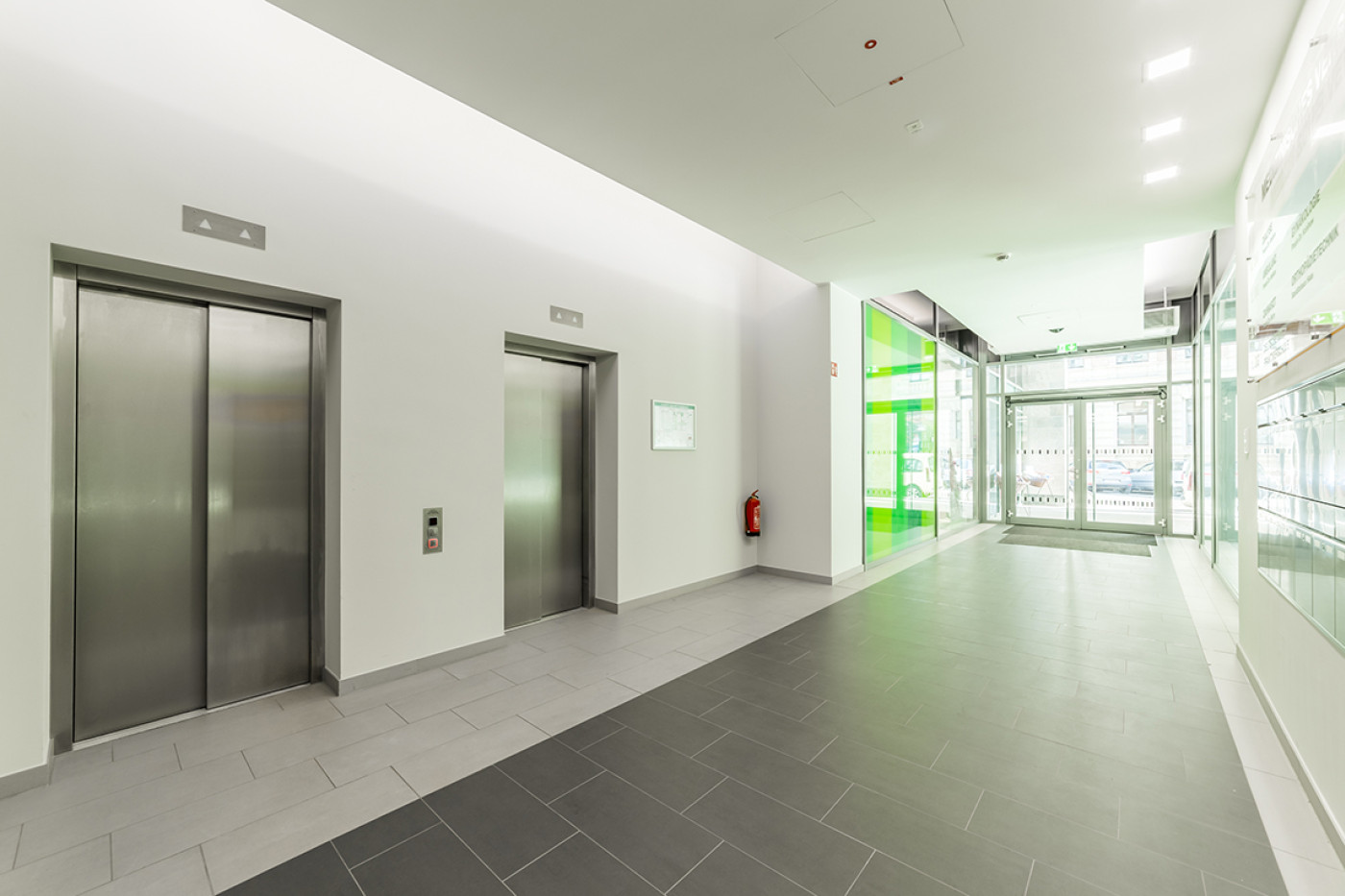 Lift lobby with two stainless steel lifts, a green-backed glass wall on the right.