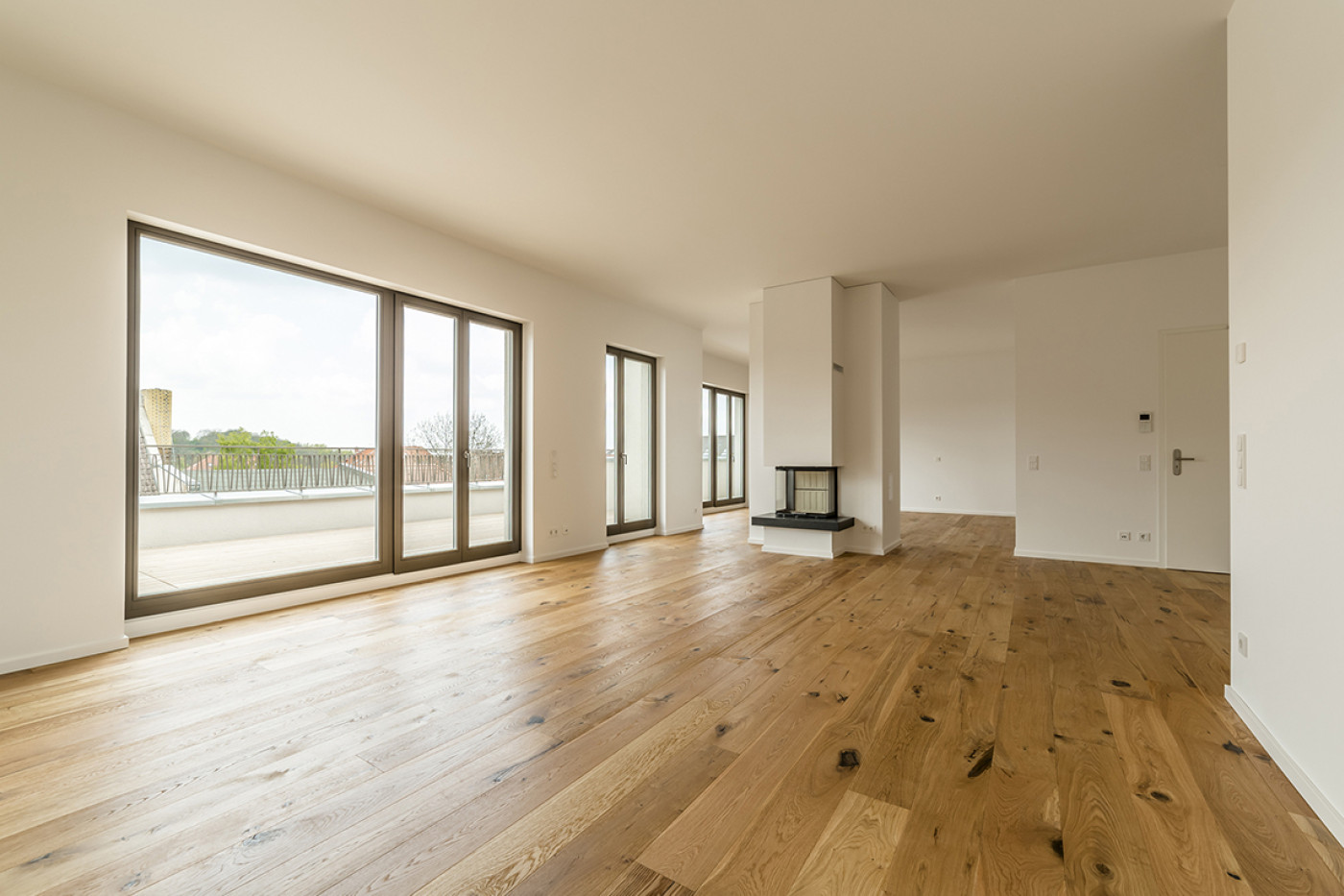 Empty living room with oak boards, a wide window wall onto the terrace and a free-standing wood stove.
