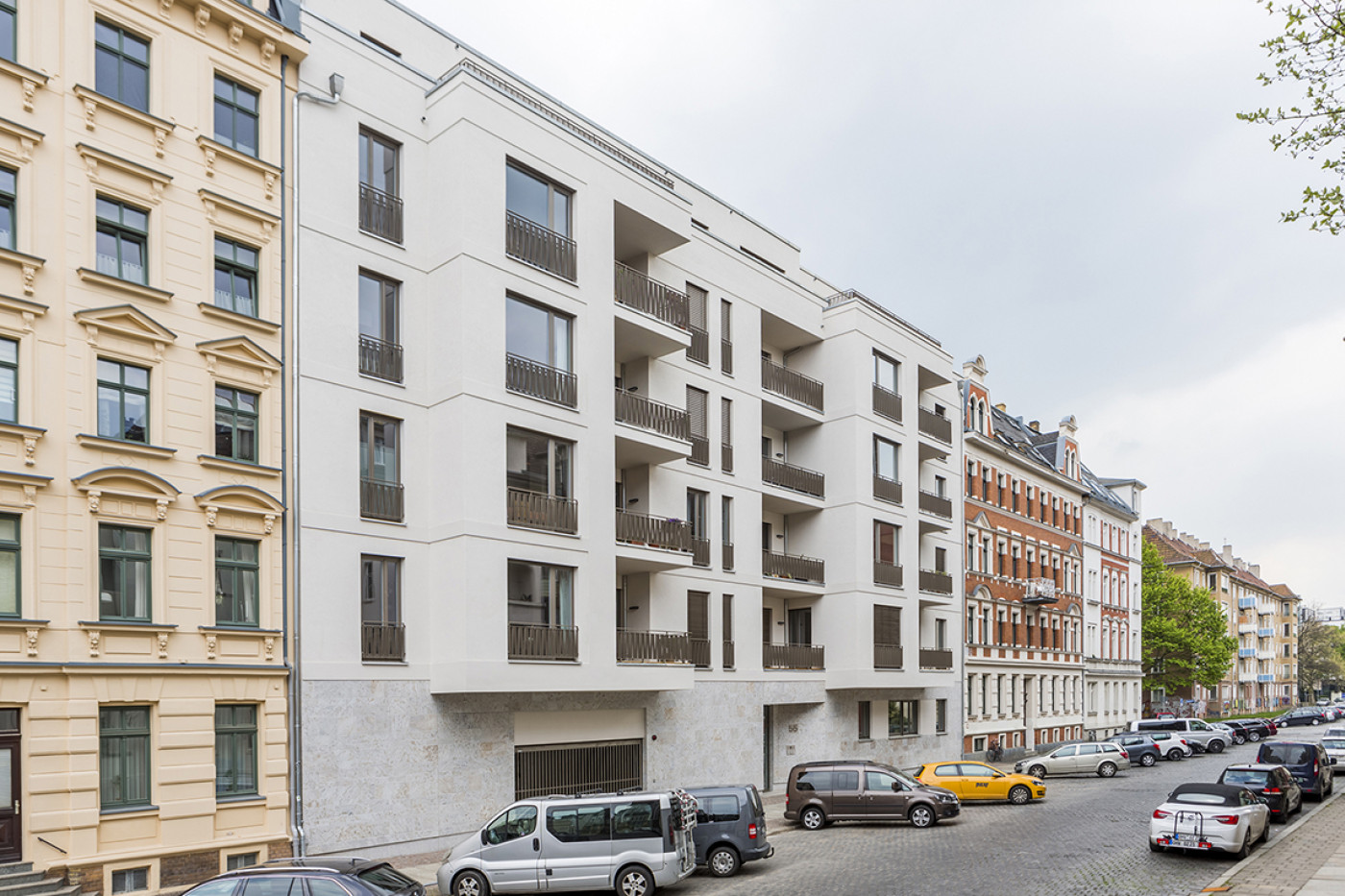 The new building in pale render with loggias in the terrace between two Gr&uuml;nderzeit buildings, parked cars in front.