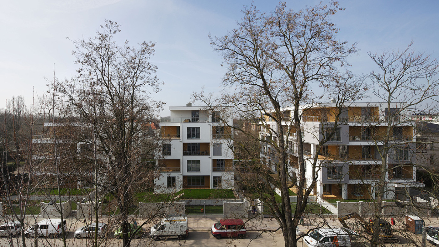 White apartment buildings with timber balconies, seen through bare trees from the opposite side of the street.